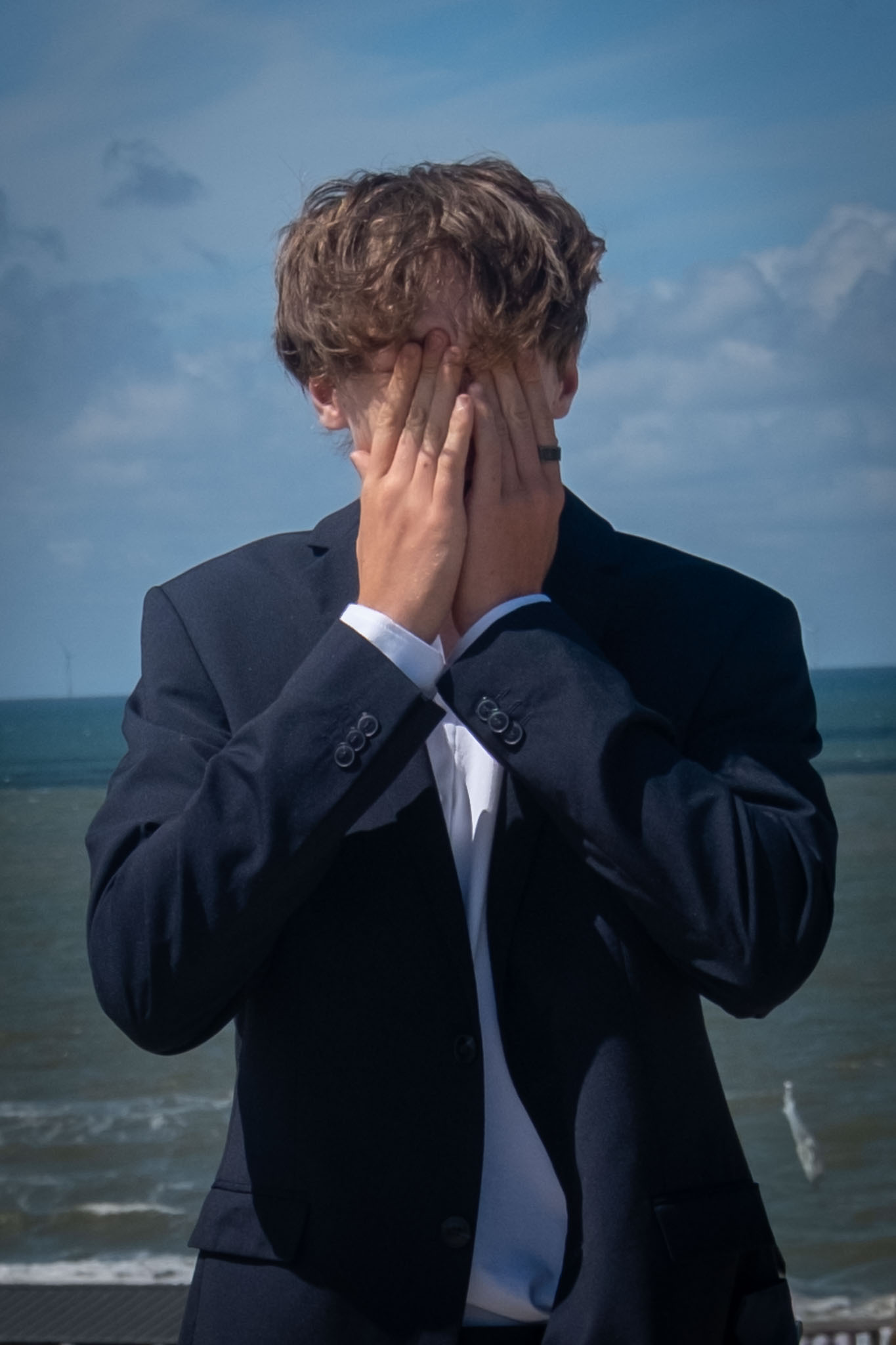 Een man in een donker pak houdt zijn handen voor zijn gezicht op het strand