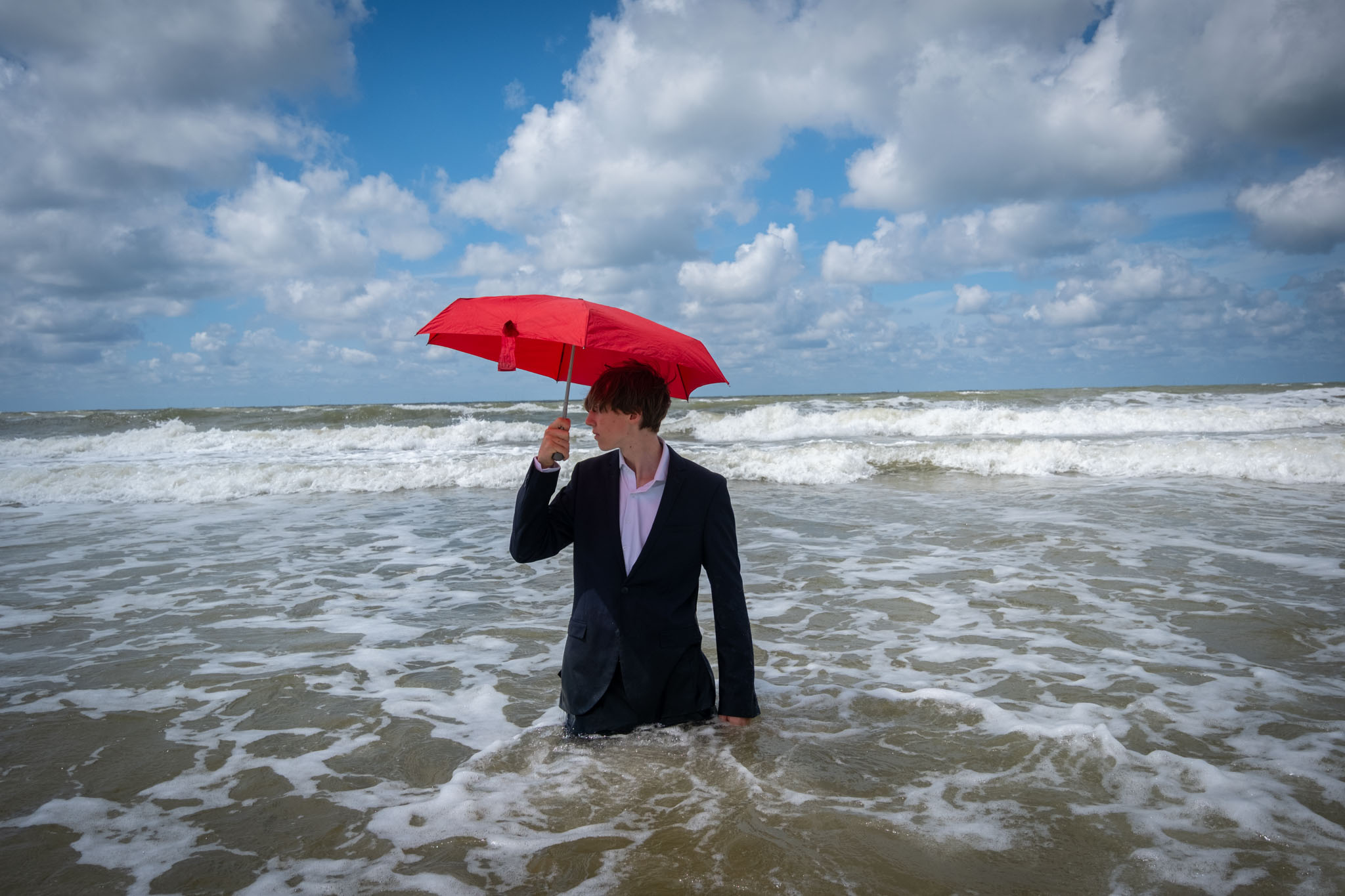 Een man in een donker pak staat met een rode paraplu in zee