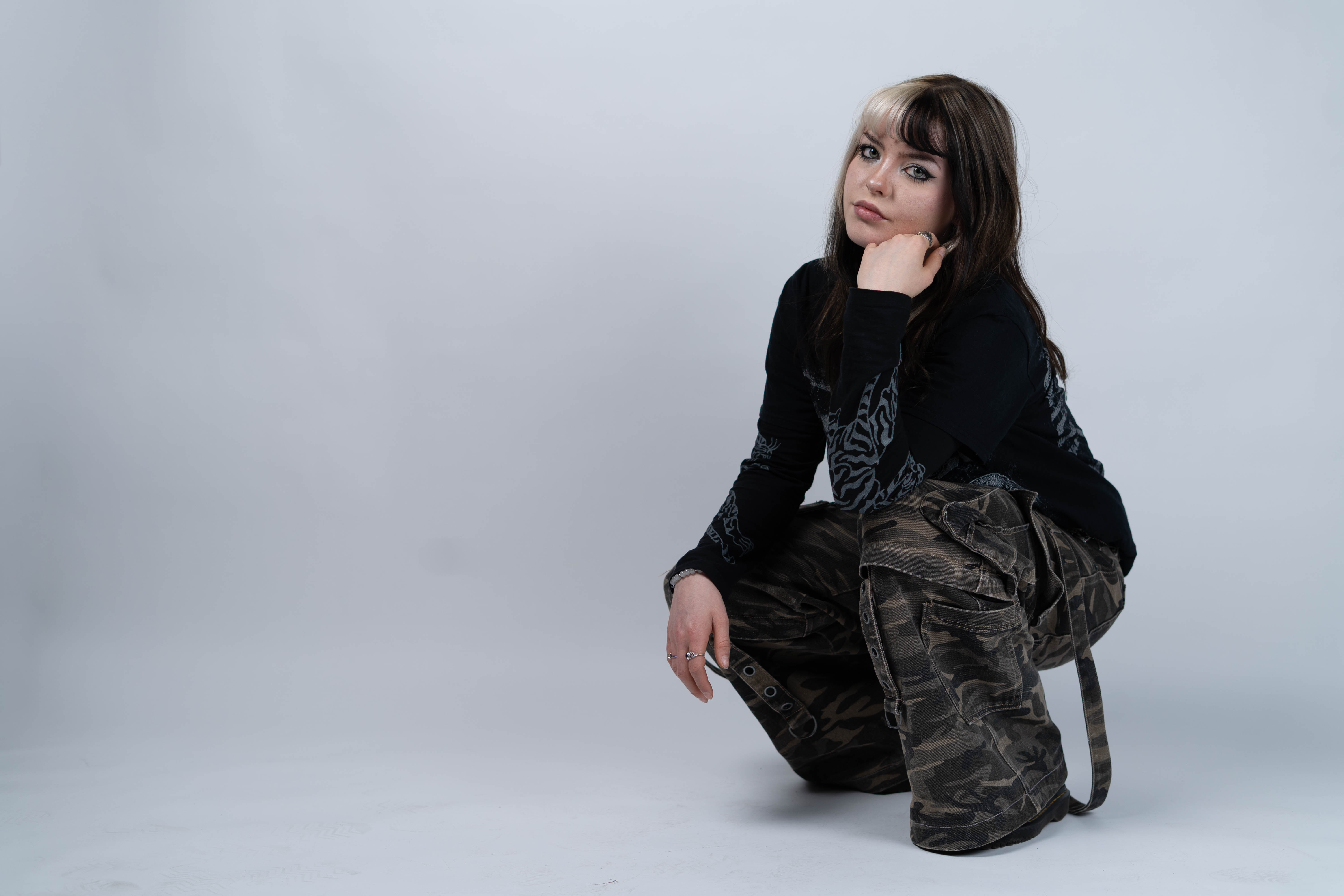 Een vrouw in donkere kleding zit gehurkt tegen een lichte studioachtergrond